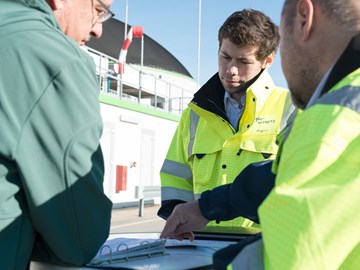 Drei Personen im Gespräch über Unterlagen vor einem Industriegebäude Drei Personen im Gespräch über Unterlagen vor einem Industriegebäude