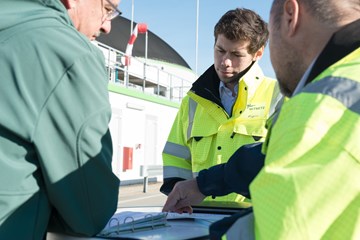 Drei Personen im Gespräch über Unterlagen vor einem Industriegebäude Drei Personen im Gespräch über Unterlagen vor einem Industriegebäude