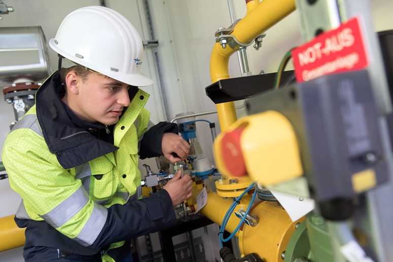 Mitarbeiter in Warnschutzjacke mit weißem Helm arbeitet an einer Anlage mit gelben Rohren Mitarbeiter in Warnschutzjacke mit weißem Helm arbeitet an einer Anlage mit gelben Rohren
