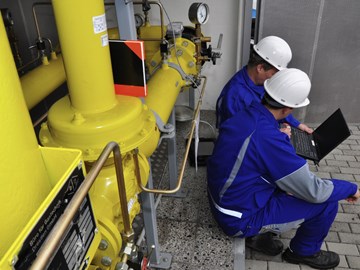 Zwei Mitarbeiter in blauer Arbeitskleidung sitzen am Fuße einer Anlage über einen Laptop gebeugt Zwei Mitarbeiter in blauer Arbeitskleidung sitzen am Fuße einer Anlage über einen Laptop gebeugt