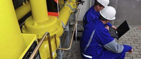 Zwei Mitarbeiter in blauer Arbeitskleidung sitzen am Fuße einer Anlage über einen Laptop gebeugt Zwei Mitarbeiter in blauer Arbeitskleidung sitzen am Fuße einer Anlage über einen Laptop gebeugt