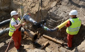 Zwei Mitarbeiter in einer Baugrube mit Rohrteilen, die gerade von einem Kran herabgelassen werden Zwei Mitarbeiter in einer Baugrube mit Rohrteilen, die gerade von einem Kran herabgelassen werden
