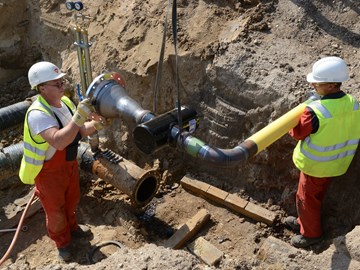 Zwei Mitarbeiter in einer Baugrube mit Rohrteilen, die gerade von einem Kran herabgelassen werden Zwei Mitarbeiter in einer Baugrube mit Rohrteilen, die gerade von einem Kran herabgelassen werden