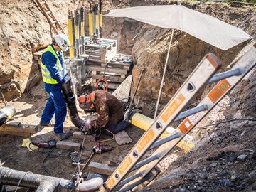 Zwei Mitarbeiter schweißen in einer Baugrube unter einem Schirm an Rohren Zwei Mitarbeiter schweißen in einer Baugrube unter einem Schirm an Rohren