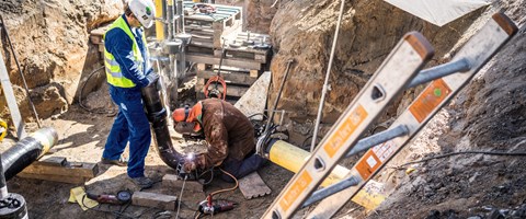 Zwei Mitarbeiter schweißen in einer Baugrube unter einem Schirm an Rohren Zwei Mitarbeiter schweißen in einer Baugrube unter einem Schirm an Rohren