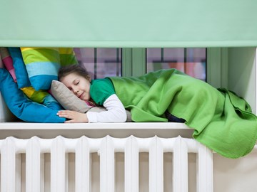 Kleines Mädchen schläft auf bunten Kissen, unter einer grünen Decke, auf dem Fenstersims über einer Heizung Kleines Mädchen schläft auf bunten Kissen, unter einer grünen Decke, auf dem Fenstersims über einer Heizung