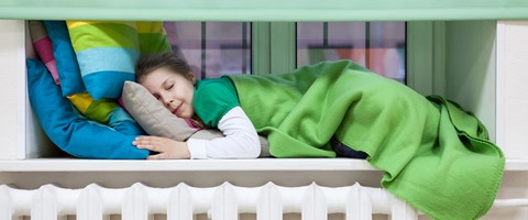 Kleines Mädchen schläft auf bunten Kissen, unter einer grünen Decke, auf dem Fenstersims über einer Heizung Kleines Mädchen schläft auf bunten Kissen, unter einer grünen Decke, auf dem Fenstersims über einer Heizung