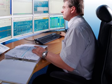 Mitarbeiter am Schreibtisch mit Unterlagen vor mehreren Monitoren Mitarbeiter am Schreibtisch mit Unterlagen vor mehreren Monitoren