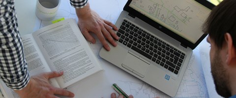 Tisch mit Laptop, Plänen, offenem Buch und Händen zweier Männer - Sicht von oben Tisch mit Laptop, Plänen, offenem Buch und Händen zweier Männer - Sicht von oben