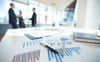 Auf einem Tisch liegen Kugelschreiber auf Diagrammen, im Hintergrund stehen drei Personen in Anzügen Auf einem Tisch liegen Kugelschreiber auf Diagrammen, im Hintergrund stehen drei Personen in Anzügen