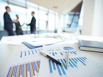 Auf einem Tisch liegen Kugelschreiber auf Diagrammen, im Hintergrund stehen drei Personen in Anzügen Auf einem Tisch liegen Kugelschreiber auf Diagrammen, im Hintergrund stehen drei Personen in Anzügen