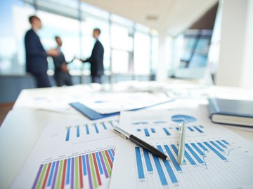 Auf einem Tisch liegen Kugelschreiber auf Diagrammen, im Hintergrund stehen drei Personen in Anzügen Auf einem Tisch liegen Kugelschreiber auf Diagrammen, im Hintergrund stehen drei Personen in Anzügen