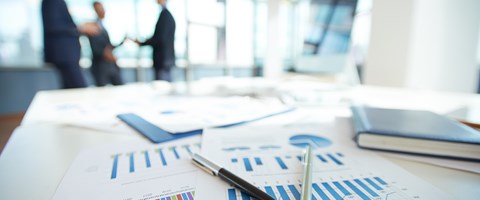 Auf einem Tisch liegen Kugelschreiber auf Diagrammen, im Hintergrund stehen drei Personen in Anzügen Auf einem Tisch liegen Kugelschreiber auf Diagrammen, im Hintergrund stehen drei Personen in Anzügen