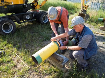 Zwei Mitarbeiter arbeiten vor einem Bagger an einer Rohrleitung Zwei Mitarbeiter arbeiten vor einem Bagger an einer Rohrleitung